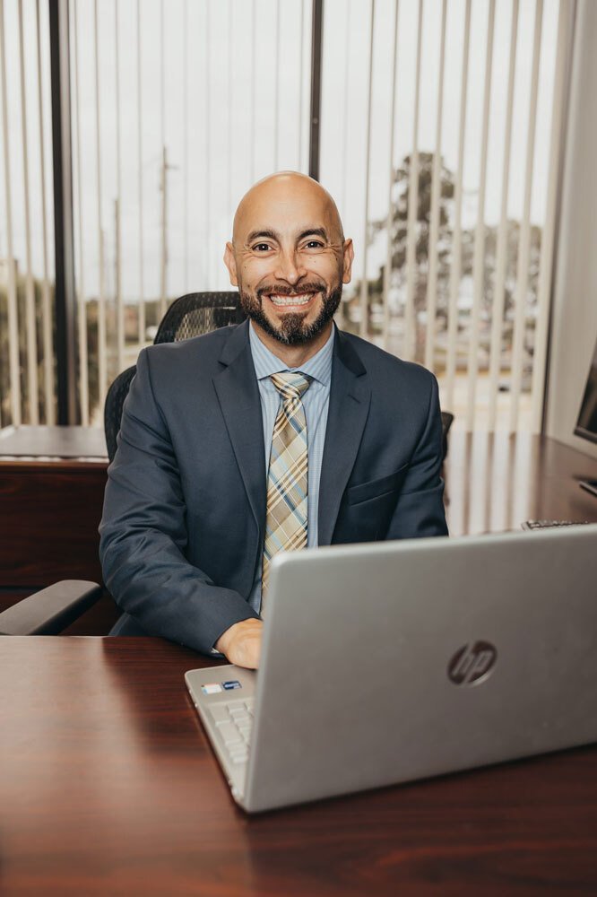 A bald man with a beard, wearing a dark blue suit and striped tie, sits at a wooden desk in an office setting, smiling at the camera. He is working on an HP laptop, with large windows and vertical blinds in the background.